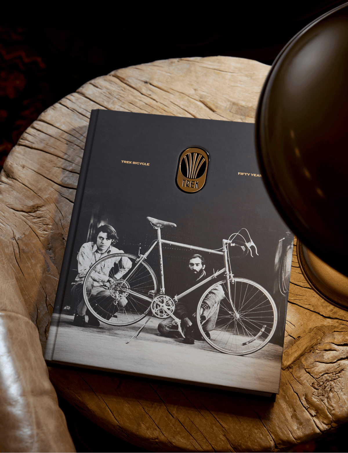 The Trek 50th Anniversary Book, a black hardcover with a black-and-white photo of two people and an early Trek bicycle, resting on a wood table