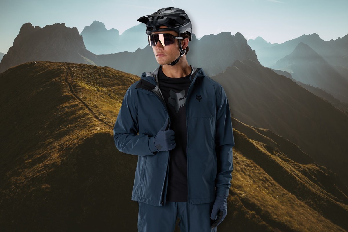 A cyclist wearing a Fox Racing jacket, gloves, helmet, and sunglasses stands on a mountain ridge at sunrise, with layered peaks in the background.