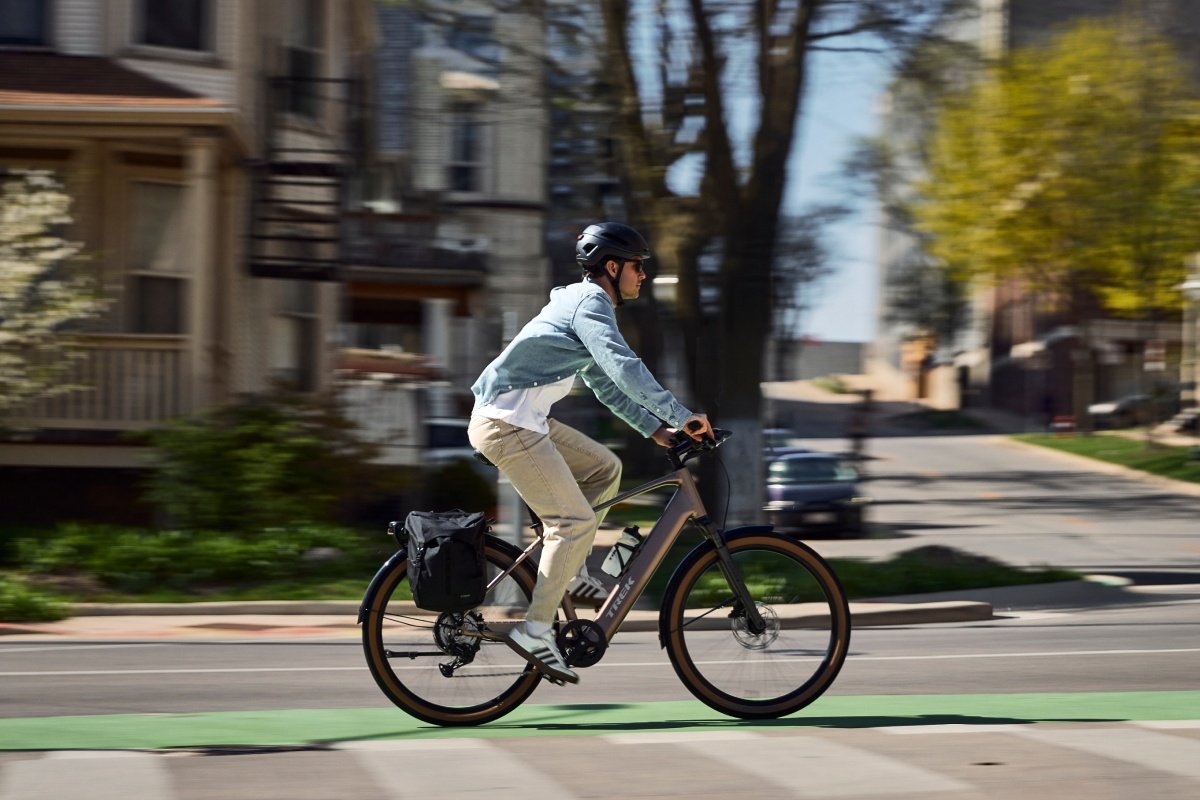 A rider in a denim jacket rides a Trek city bike with a rear pannier down a tree-lined street.