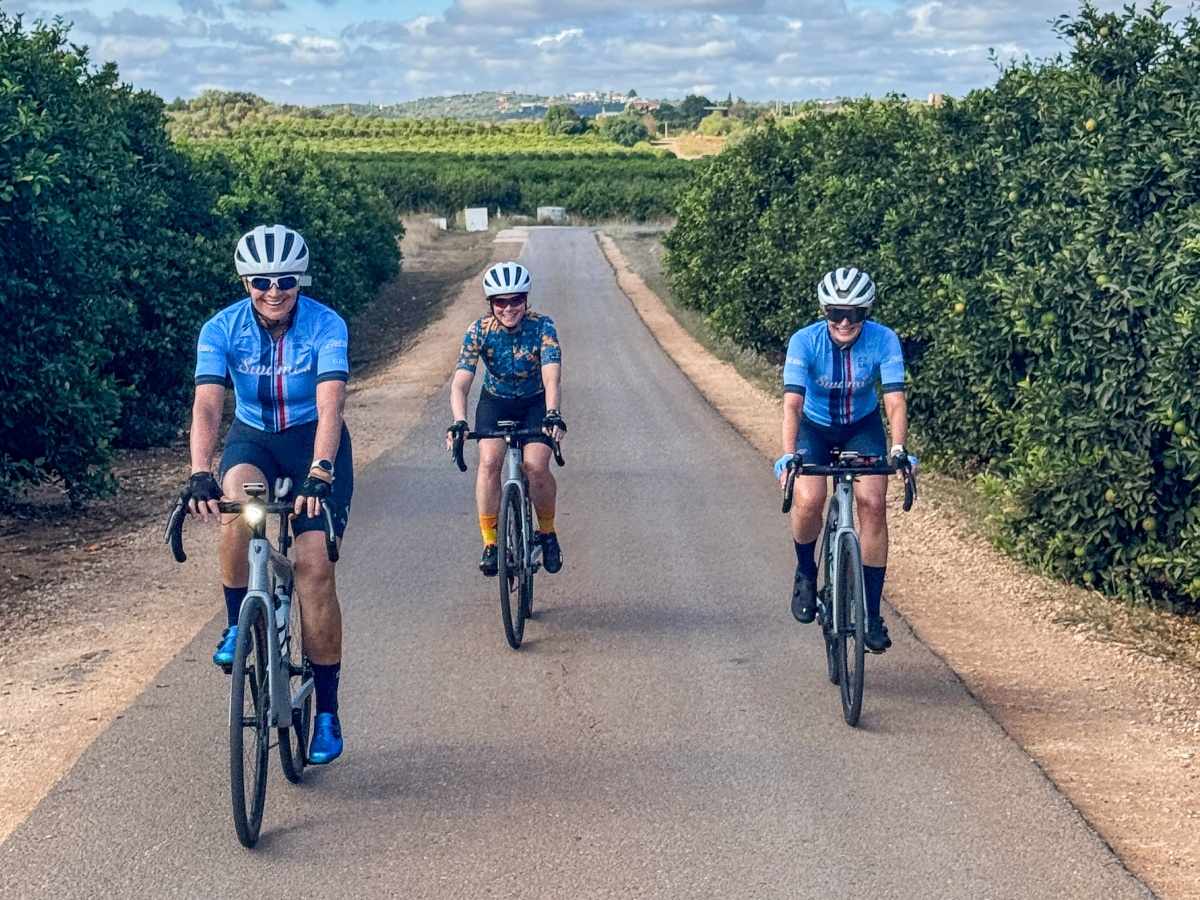 Three cyclists in blue kits riding a quiet road between orange groves in Portugal