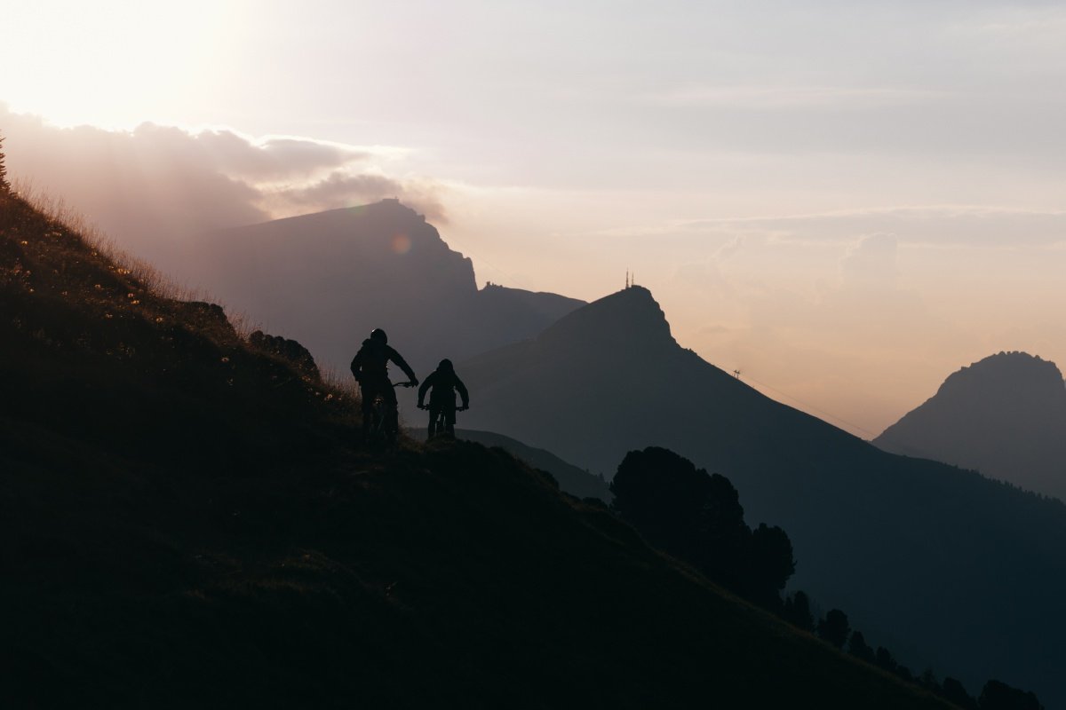 Two mountain bikers silhouetted on a hillside at sunset with mountains behind
